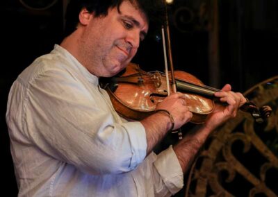 Man with dark curly hair playing violin in white shirt against dim background with decorative ironwork
