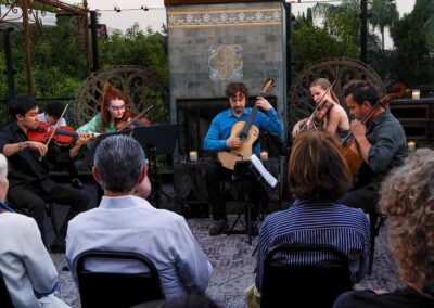 Chamber music ensemble with violins, guitar, and cello performing for an intimate audience at an outdoor evening concert.