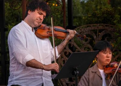 Two musicians playing violins during an outdoor classical music performance in a garden setting with ornate metalwork.