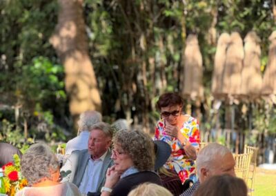 Group of older adults socializing at an outdoor garden event with lush greenery and floral table decorations.