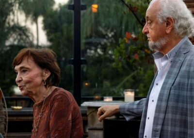 Elderly man and woman standing outdoors at an evening event with greenery and candles in the background.