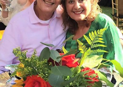 Smiling couple posing together at outdoor event table with vibrant floral centerpiece of sunflowers and red roses.