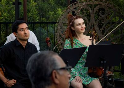 Chamber musicians with violins and music stands performing at an outdoor garden concert surrounded by greenery.