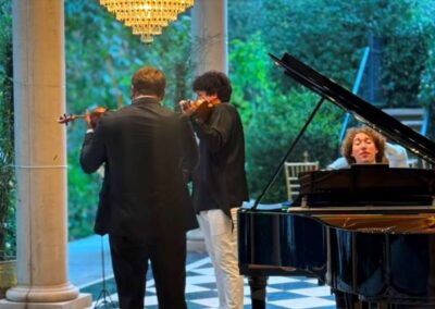 Two violinists and a pianist perform classical music under a crystal chandelier on a checkered marble floor.