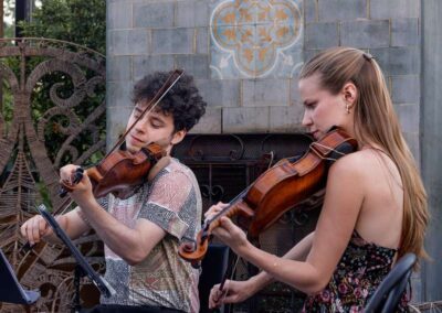 Two violinists, a man and a woman, performing outdoors near a decorative stone fireplace and ornate metalwork.