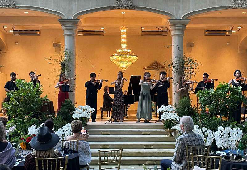 Meet the Artists Chamber orchestra of violinists performing on stage under an ornate chandelier at an elegant indoor concert venue.