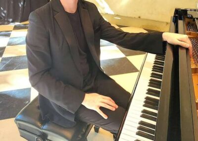 Smiling young man with curly hair in black suit sitting at a grand piano on a checkered floor.