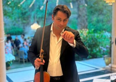 Male violinist in black suit holding violin and bow at elegant event with chandelier and string lights.