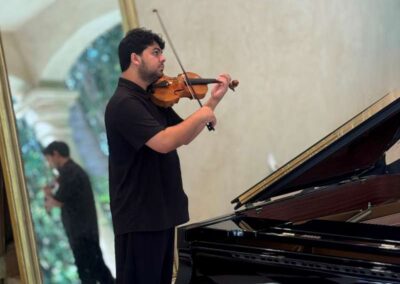Violinist in black attire playing violin beside a Steinway & Sons grand piano near an ornate gold mirror.