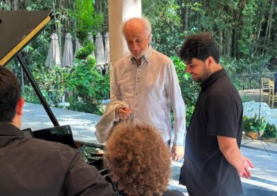 Elderly music instructor teaching students at a grand piano in an elegant garden venue with lush greenery.
