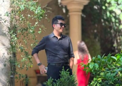 Man in black shirt and sunglasses holding phone stands among white orchids and greenery in an elegant garden patio.