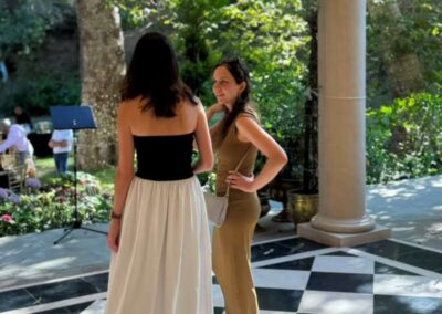 Two women in elegant dresses chatting on a black and white checkered patio surrounded by lush green garden trees.
