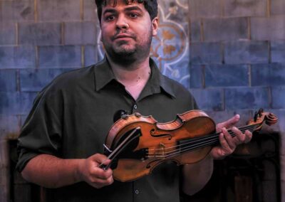Man in dark shirt holding a violin and bow against a decorative blue-tiled wall in a concert setting.
