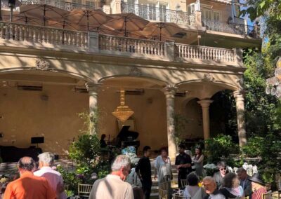 Elegant outdoor garden party at a Mediterranean-style mansion with guests, dining tables, chandelier, and grand piano.