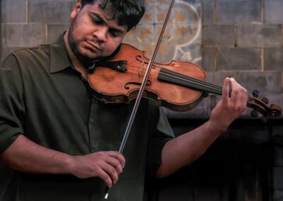 Violinist in dark green shirt performing on stage against a stone wall with decorative tile detail.
