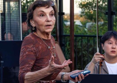 Elderly woman speaking at outdoor event holding phone, with violinist playing behind her at dusk under a pergola.
