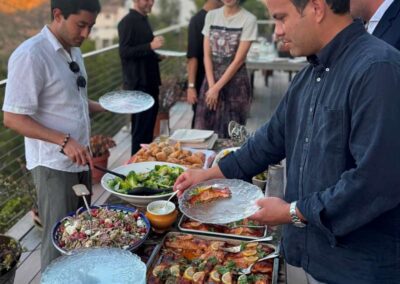 Guests serve food from an outdoor buffet on a hillside patio at sunset, with salads, meats, and braided bread.