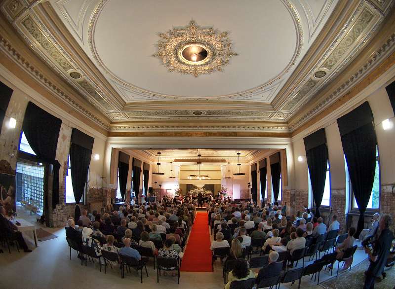 Marienbad concert. Audience seated in ornate historic concert hall with domed ceiling and red carpet aisle watching a live performance on stage