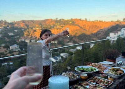 Woman raising a glass in a toast at a hilltop dinner party with a food-laden table at golden hour sunset.