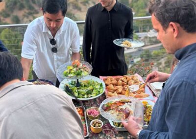 Men serving food from buffet table at outdoor hillside gathering with scenic canyon view at sunset.