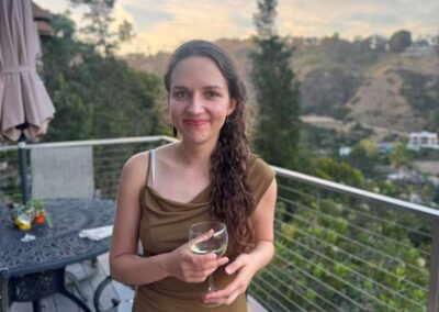 Woman in olive dress holding white wine on hillside deck at sunset with scenic canyon view.