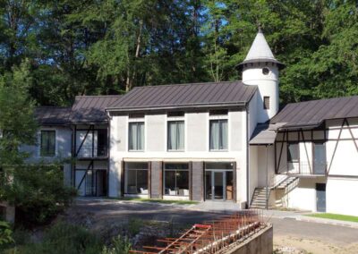 Modern white building with dark metal roof and conical turret tower nestled among lush green forest trees.