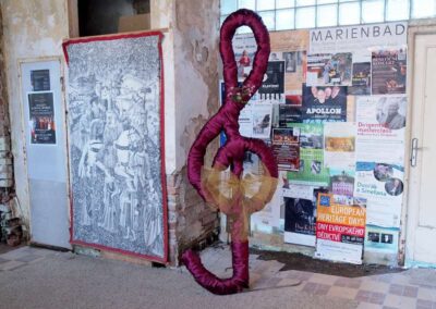 Giant burgundy fabric treble clef sculpture in weathered room with MARIENBAD concert posters and tapestry
