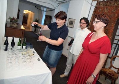 Woman pouring red wine into glass at wine tasting event with two guests observing in elegant interior setting
