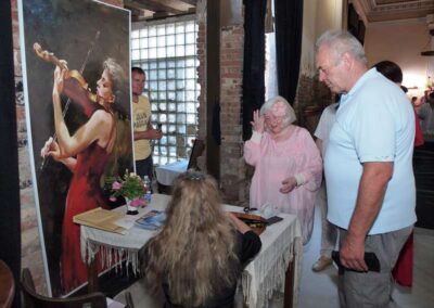 Elderly woman in pink dress speaks animatedly at art exhibition beside violinist painting and gathered visitors.