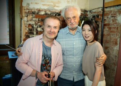 Three musicians posing together, one holding a violin and bow, in front of an exposed brick wall backstage.