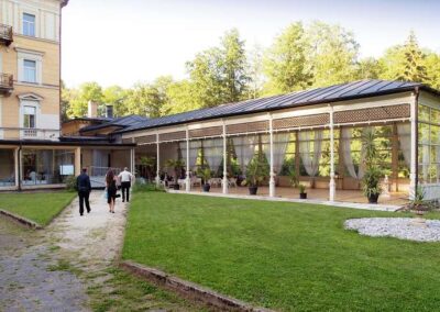 Elegant hotel terrace pavilion with white curtains and columns, guests walking on garden path surrounded by lush greenery