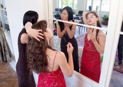 Women in elegant dresses getting ready and styling hair in front of a large mirror before an event
