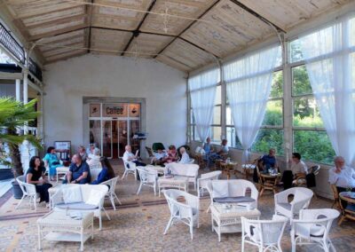 Guests relaxing in vintage white wicker chairs at an old-world conservatory café with large windows and sheer curtains.