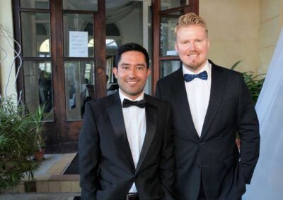 Two smiling men in black tuxedos with bow ties standing together at a formal event near glass doors.