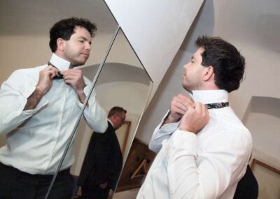 Man in white dress shirt adjusting bow tie while looking in mirror, getting ready for formal event