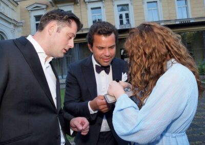 Two men in formal tuxedos and woman in blue dress viewing smartphone outside elegant historic building