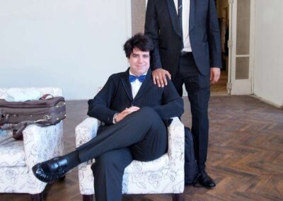 Two men in formal suits pose in a bright room, one seated in a floral armchair, the other standing beside him.