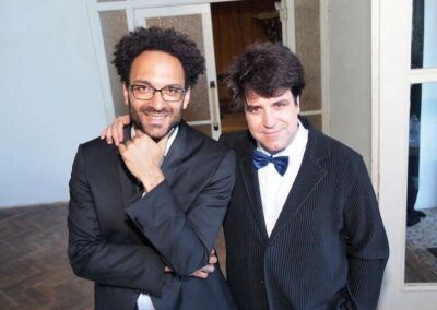 Two smiling men in formal black suits posing together, one wearing glasses and one wearing a navy bow tie at an event.
