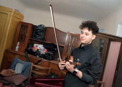 Young male violinist in black shirt holding violin and bow, standing in room with vintage wooden cabinets