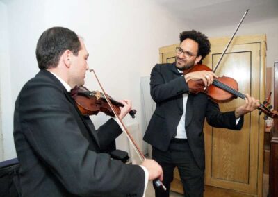 Two men in formal black suits playing violins together in a room, one smiling while performing.