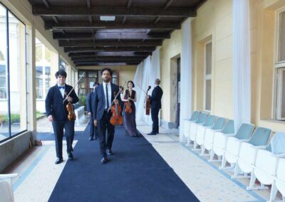 Classical string quartet musicians in formal attire walking along outdoor covered terrace carrying violins and cello