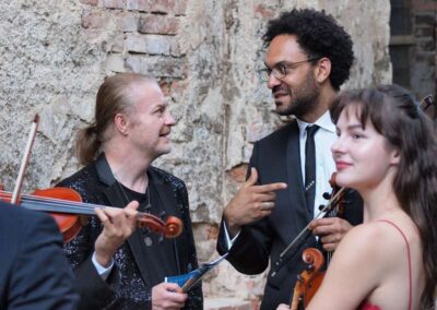 Three classical musicians with violins conversing against a rustic brick wall backdrop at an outdoor event.