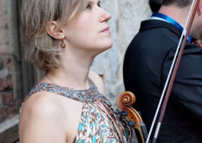 Woman in patterned dress holding violin and bow, gazing upward at outdoor musical event with musician nearby