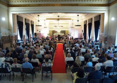 Audience seated in an ornate concert hall watching an orchestra perform on stage with a red carpet aisle.