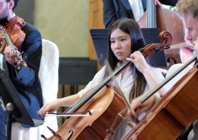 Young female cellist performing with orchestra, focused expression while playing cello among string musicians in concert.