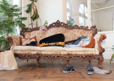 Ornate carved wood Victorian sofa with floral upholstery holding a guitar case, near sandals on parquet floor.
