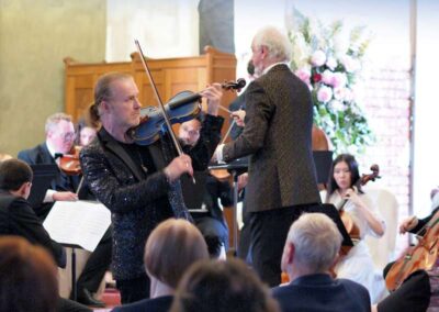 Violin soloist in sparkling jacket performs with conductor and orchestra during a classical concert.