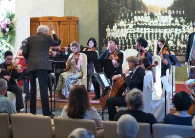 Chamber orchestra performing with conductor leading string musicians on violins, violas, and cellos before an audience.