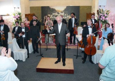 Elderly conductor on wooden platform acknowledging applause with orchestra musicians standing behind him at concert venue