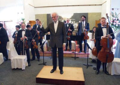 Elderly orchestra conductor standing on podium with musicians holding violins and cellos at formal concert performance.